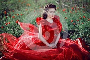 Beautiful blue-eyed young lady with perfect make up and hairstyle wearing luxurious silk red ball gown sitting in the poppy field