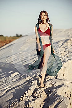 beautiful boho girl in red swimsuit walking on sandy beach at sunset light. attractive young woman in bohemian look with win