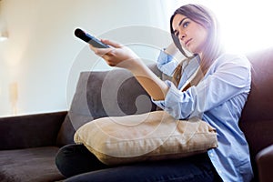 Beautiful bored young woman watching TV and holding remote control at home.
