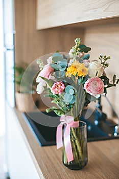 A beautiful bouquet of assorted flowers placed in a clear glass vase with a pink ribbon, set on a countertop against the backdrop