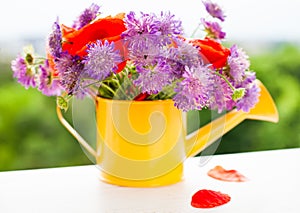 Beautiful bouquet of bright wildflowers in yellow watering can and two fallen petals of poppy on white wooden background. Close-up