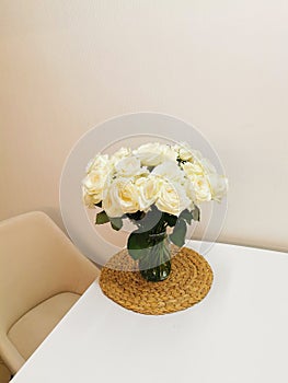 A beautiful bouquet of white roses in a glass vase on a white table and interior on a wicker placemat. Minimalistic
