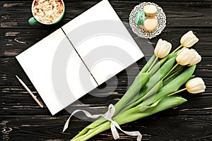 Beautiful bouquet of white tulips, empty open notebook, silver pen, cup of cappuccino, makaroons on the black wooden background