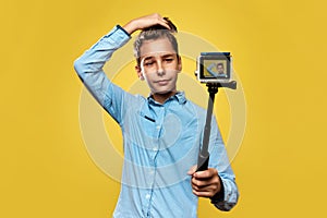 Beautiful boy blogger makes a selfie or video on a compact camera with the monopod. Yellow background. Copy space