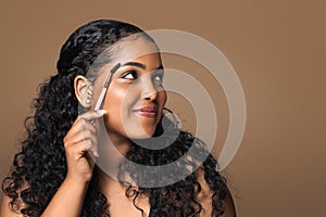 Beautiful brazilian body positive woman brushing eyebrows holding brush, looking aside at free space, brown background