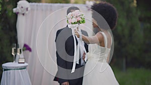 Beautiful bridal bouquet in stretched female hand with blurred happy couple of African American newlyweds talking