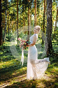 Beautiful bride with a bouquet in her hand posing on her wedding day among the green trees in summer forest. The happy bride smile