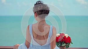 A beautiful bride with a bouquet of roses stands against the background of the sea on her wedding day