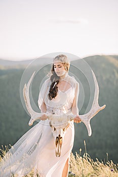 Beautiful bride brunette girl in white long dress is made of tulle looks down, keeps on hand a deer skull with horns decoration on