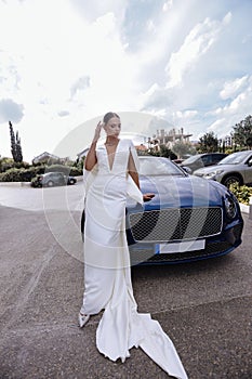beautiful bride in elegant wedding dress and accessories posing near luxury car