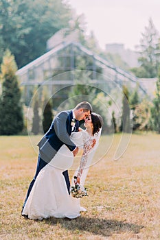 Beautiful bride with gorgeous white wedding dress and groom in stylish blue suit posing outdoor on lawn. Greenhouse at