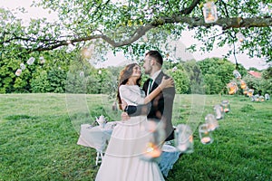 Beautiful bride and groom in evening park embracing under tree decorated with many lanterns