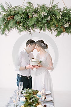 Beautiful Bride and groom look at each other at the table in the banquet hall and holding wedding cake decorated with