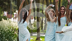 Beautiful bride and her pretty bridemaids in pale blue dresses walking in the park cheering waving hands. Woman with
