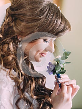 Beautiful bride with long curly hair and veil looking at the buttonhole indoors, close-up