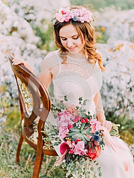 Beautiful bride is looking at the wedding bouquet while sitting on the chair in the mountains. photo