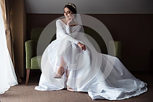 Beautiful bride in a luxurious dress, with makeup and hairstyle sitting on a sofa by the window. The bride is waiting for the