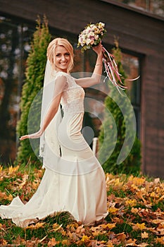 Beautiful bride in luxury white long dress with wedding bouquet is having fun and dances in autumn park on wedding day