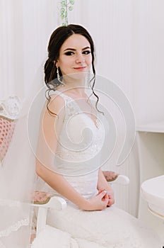 Beautiful bride perfect style. Wedding hairstyle make-up dress and bride`s bouquet. Young attractive bride on chair in white room