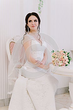 Beautiful bride perfect style. Wedding hairstyle make-up dress and bride`s bouquet. Young attractive bride on chair in white room