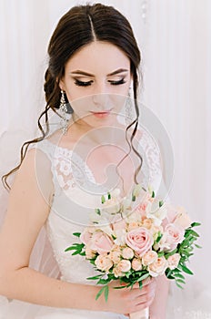 Beautiful bride perfect style. Wedding hairstyle make-up dress and bride`s bouquet. Young attractive bride on chair in white room