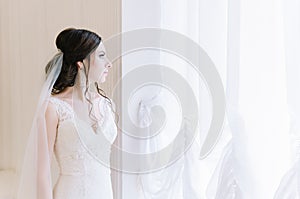 Beautiful bride perfect style. Wedding hairstyle make-up dress and bride`s bouquet. Young attractive bride near window in white r