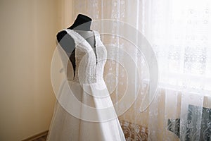 Beautiful bride`s white wedding dress hangs near the bed in a hotel room with flowers at the bottom. Bridal morning before