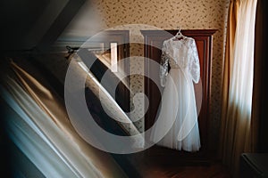 Beautiful bride`s white wedding dress hangs near the bed in a hotel room with flowers at the bottom. Bridal morning before