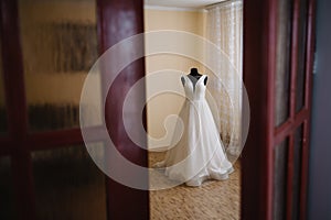 Beautiful bride's white wedding dress hangs near the bed in a hotel room with flowers at the bottom. Bridal morning