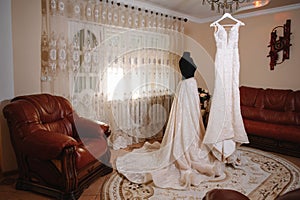 Beautiful bride's white wedding dress hangs near the bed in a hotel room with flowers at the bottom. Bridal morning