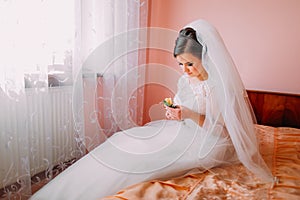 Beautiful bride sitting on a bed near the window in waiting and holding cute little wedding boutonniere