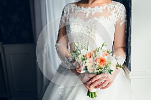 A beautiful bride is standing near the window and holding a wedding bouquet with white roses and peach peonies. Close-up