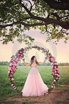 Beautiful bride standing at a round flower arch under a large single tree in a field at sunset.