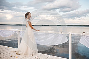 Beautiful bride stands on the pier. Water and cloudy sky on the background