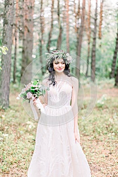 Beautiful bride walking in a coniferous forest in a wreath on her head and a luxurious wedding dress, holding rustic