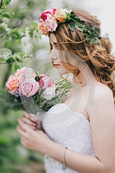 Beautiful bride in a wedding dress with bouquet and roses wreath posing in a green garden