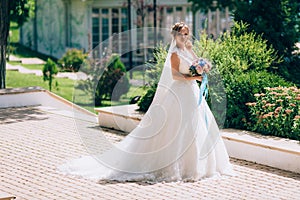 Beautiful bride in a white dress posing outdoors in a park. The girl`s bouquet is rewritten with blue ribbons.