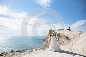 Beautiful bride in white dress stands on rock background of ocean