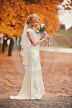 Beautiful bride in white long dress is smelling wedding bouquet in autumn park on wedding day