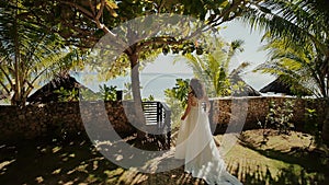 A beautiful bride in a white long wedding dress walks along a path among the palm trees to the groom. Solemn moment. The