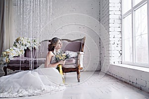 Beautiful bride in white wedding dress is sitting near sofa in front of window and holding bouquet.