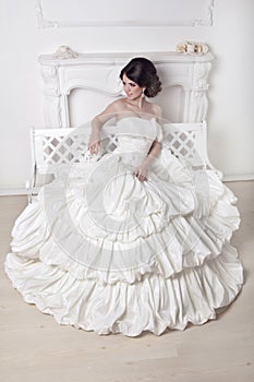 Beautiful bride woman sitting on luxuriant wedding dress over white modern interior