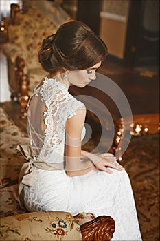 Beautiful bride, young model brunette woman, in stylish wedding dress with naked back sits on the vintage sofa and posing in luxur