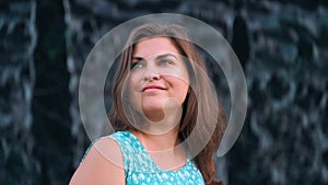 A beautiful brown-haired woman with blue eyes in a blue dress stands near the flowing water enjoys and smiles Slow motion