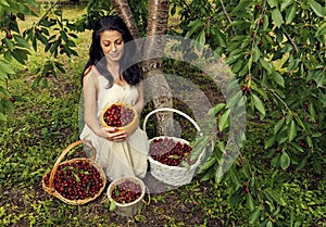Beautiful brunet woman wearing white dress under the cherry tree with two wicker baskets and two buckets full of ripe fruits