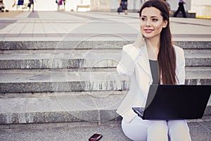 Beautiful brunette business woman in white suit with notebook on her lap, typing, working outdoors