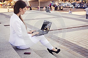 Beautiful brunette business woman in white suit with notebook on her lap, typing, working outdoors