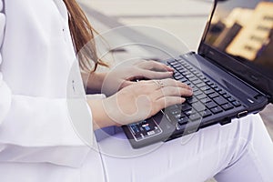 Beautiful brunette business woman in white suit with notebook on her lap, typing, working outdoors. Hands on a keyboard close up