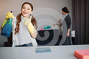 Beautiful brunette caucasian mother and daughter cleaning together in room. Girl hold spray in hand and show big thumb