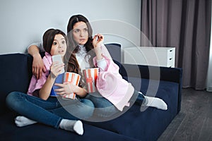 Beautiful brunette caucasian mother and daughter sit together in room. Scared adult and small women watch movie and eat
